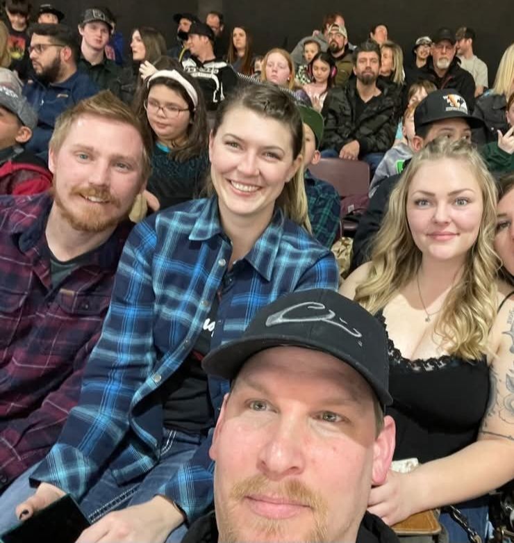 Four smiling people in the front row at an indoor event, with a crowd seated behind them.