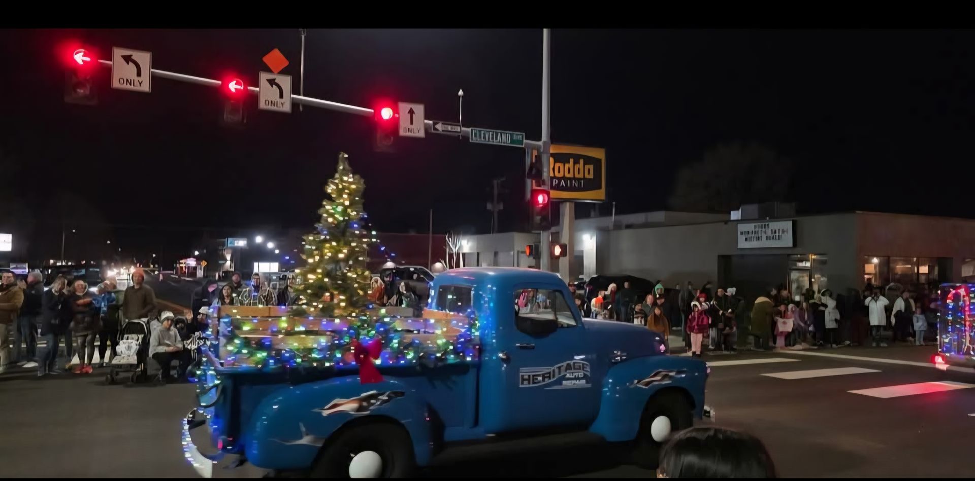 Blue vintage pickup decorated with Christmas lights drives past a crowd at a nighttime parade.