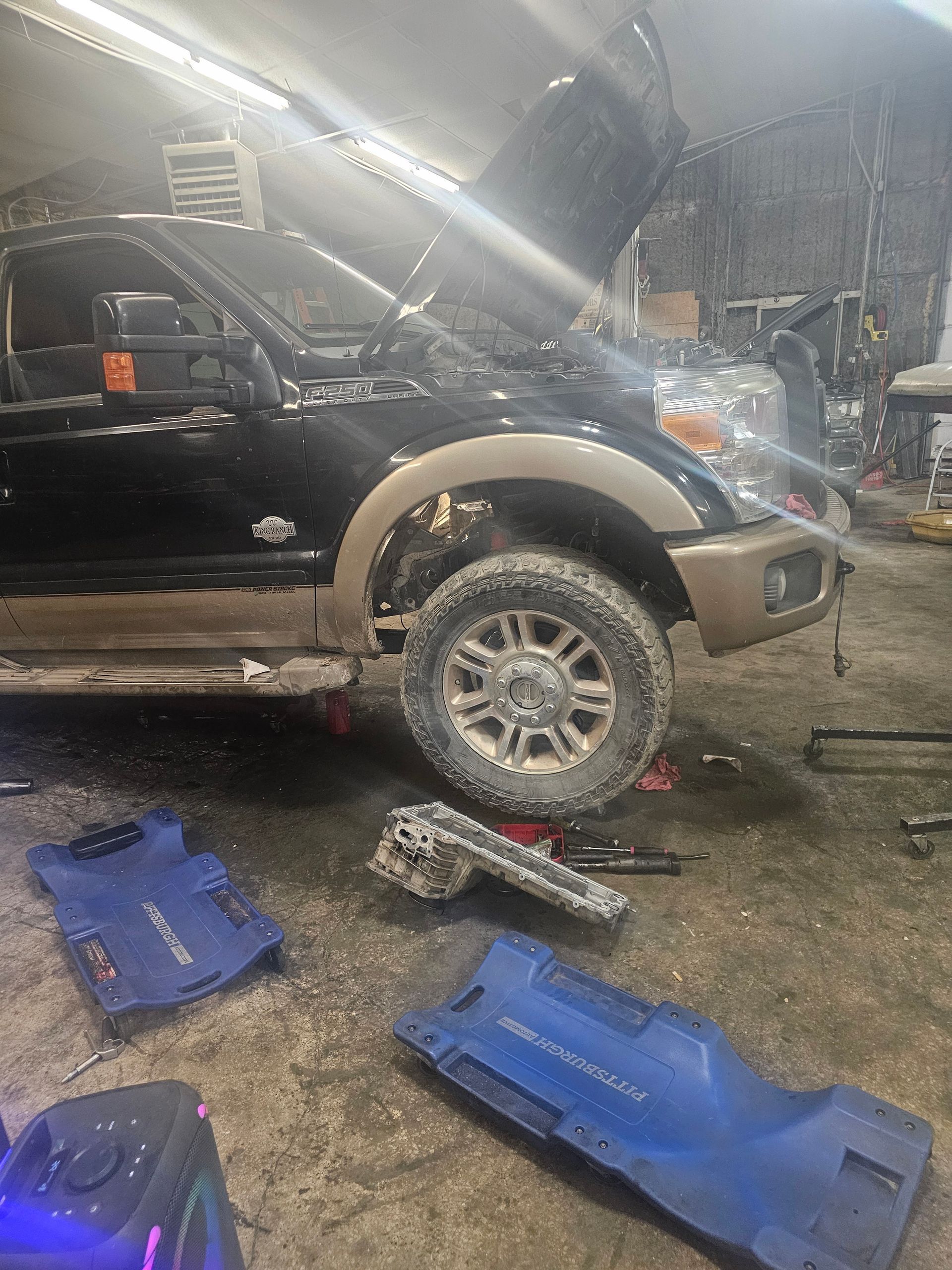 Black and tan truck with hood up, front tire removed, in a mechanic's shop. Blue creepers on the floor.