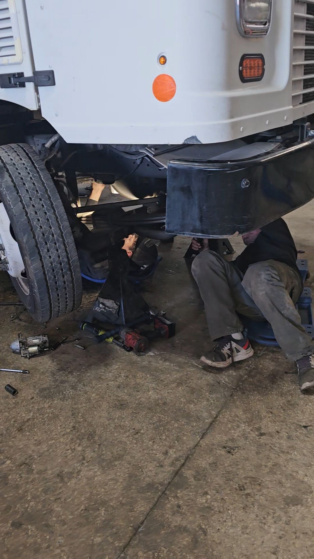 Mechanic working under a white truck, likely repairing it. Tools and parts are scattered on the garage floor.
