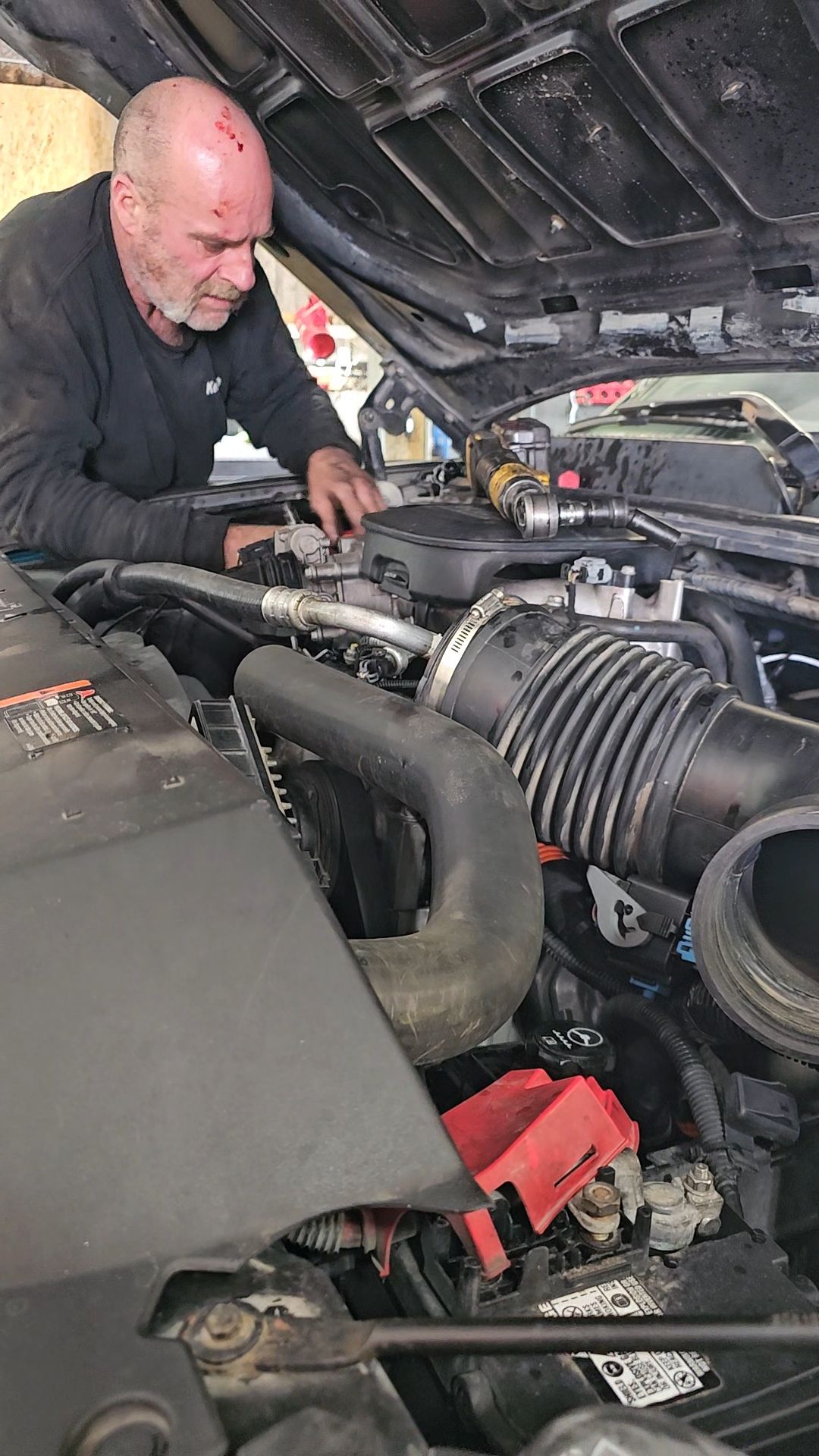 Mechanic working on a car engine under the hood. Black shirt, hands working, engine components visible.