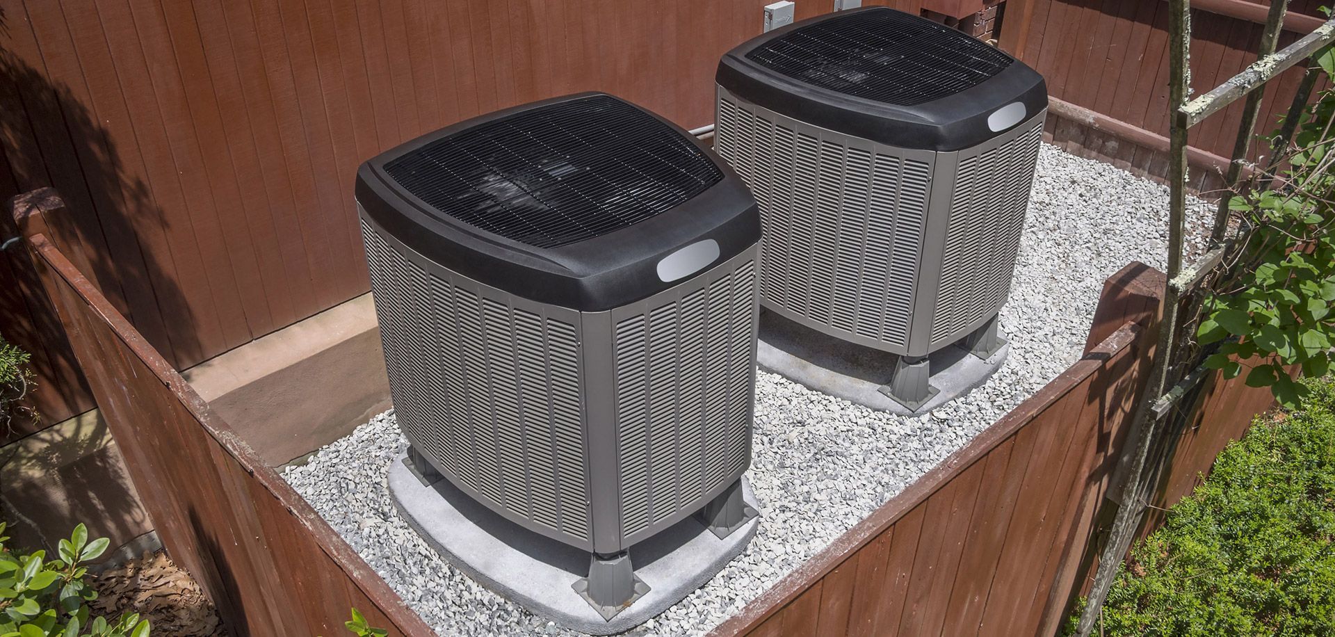 two gray outdoor air conditioning units on a gravel bed beside a wooden fence