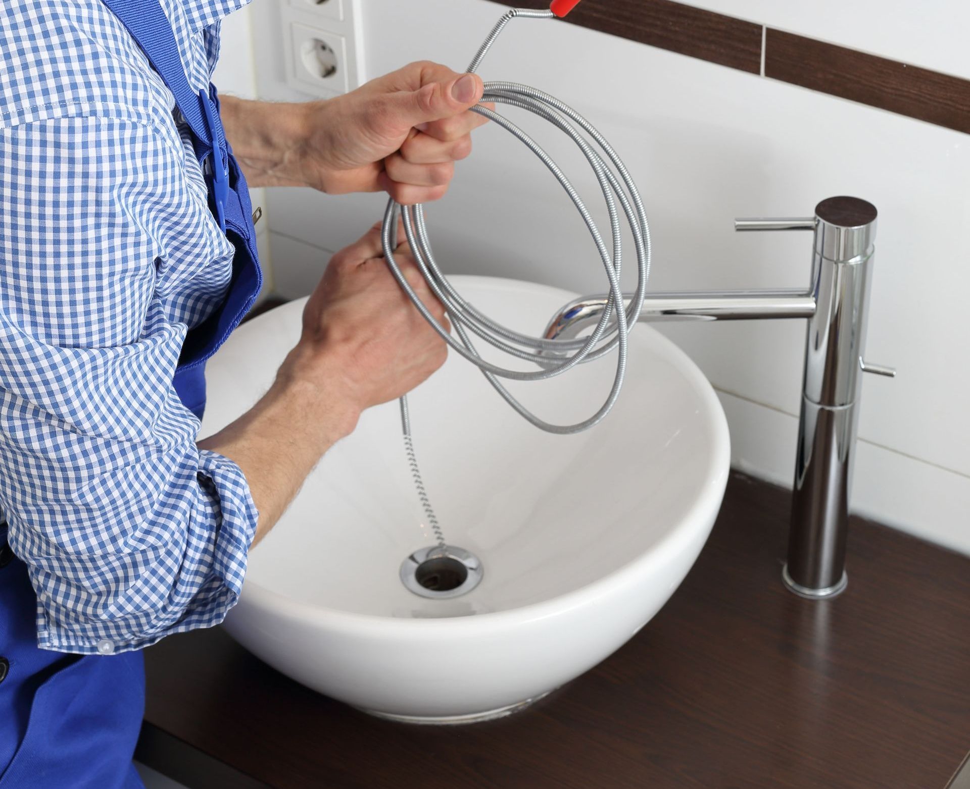 A plumber unclogs a bathroom sink with a drain snake. He wears a blue jumpsuit and holds the coiled tool.