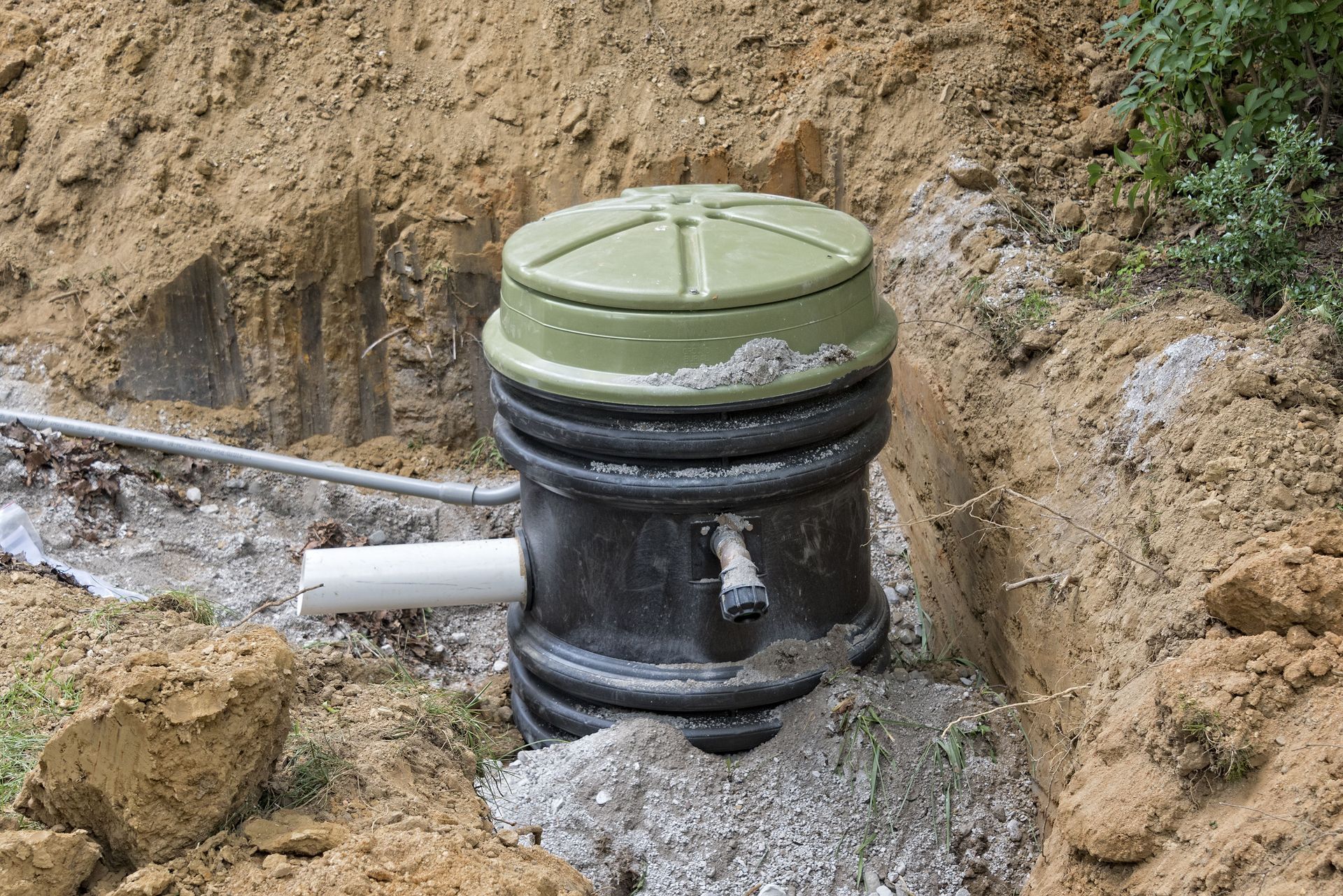 Black and green septic tank in a dirt trench, with piping and a partially exposed lid. The surrounding soil is brown and grey.