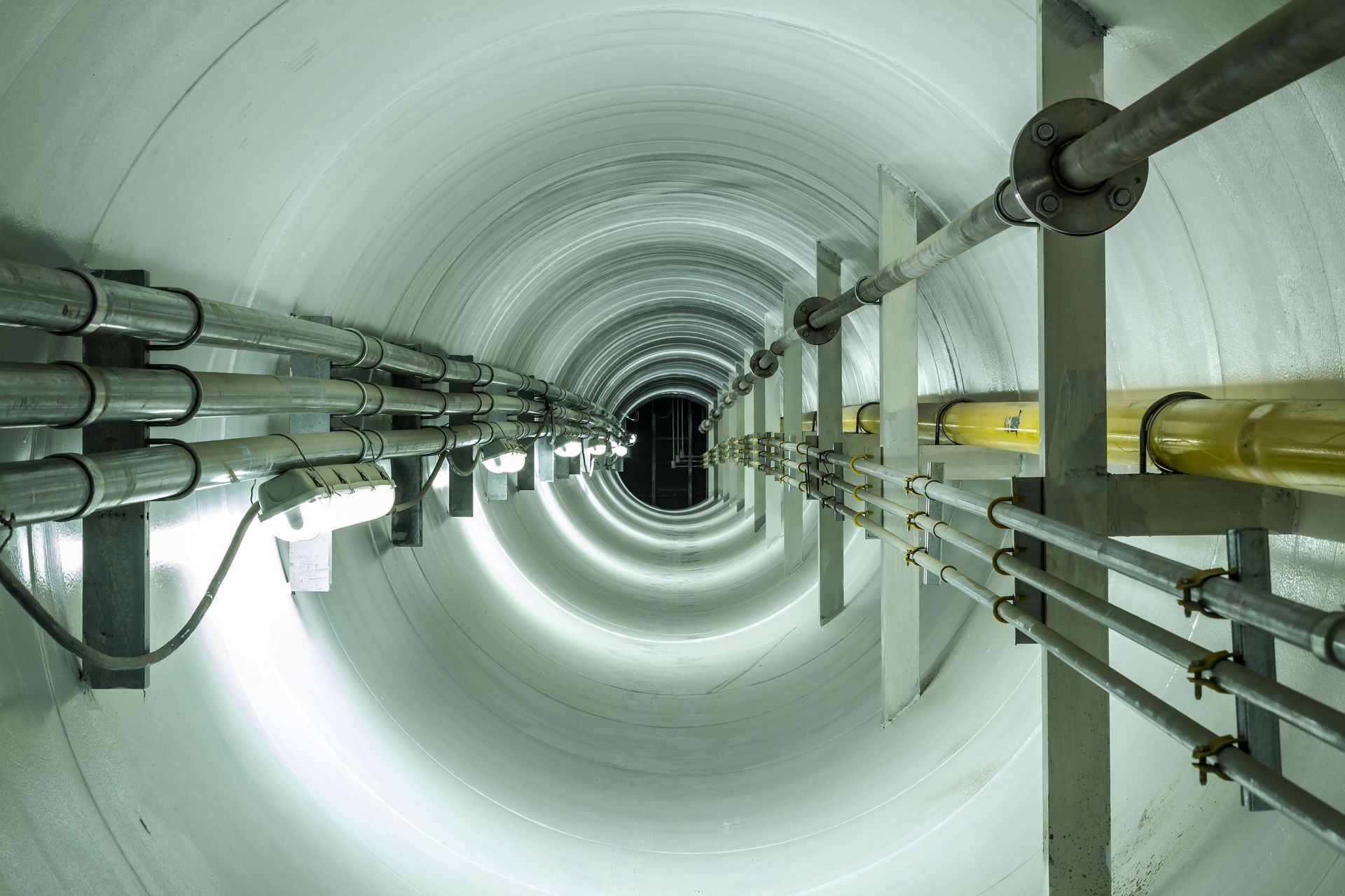 Inside of a long, white cylindrical tunnel with pipes and cables lining the walls.
