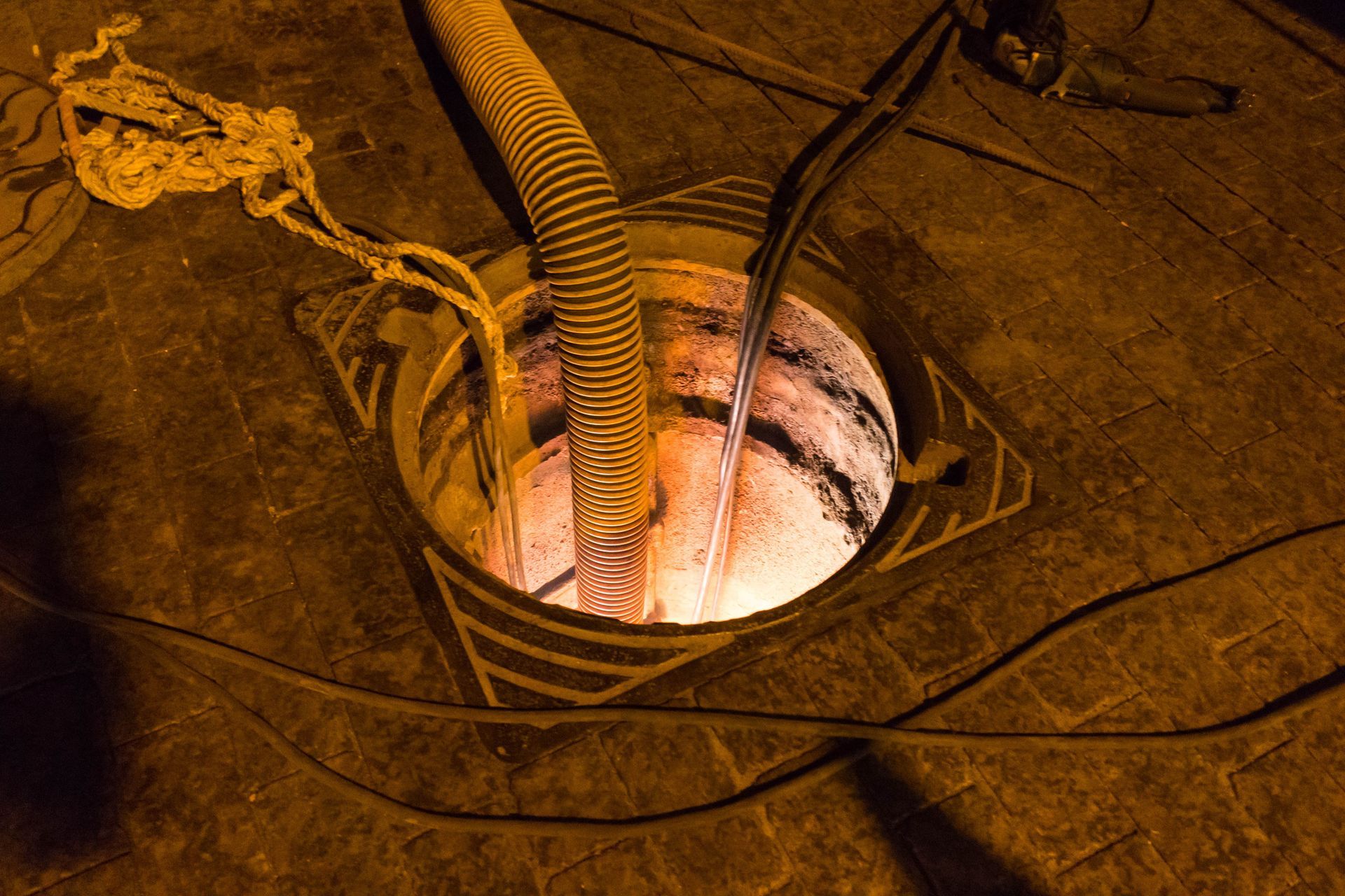 Looking down into an open manhole, electrical cables and a flexible conduit are visible. The scene is lit with warm colors.