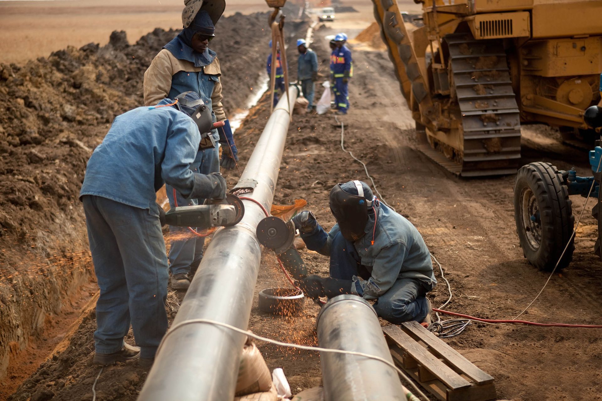 Pipeline welders working on a pipe in an open trench, with heavy machinery nearby.