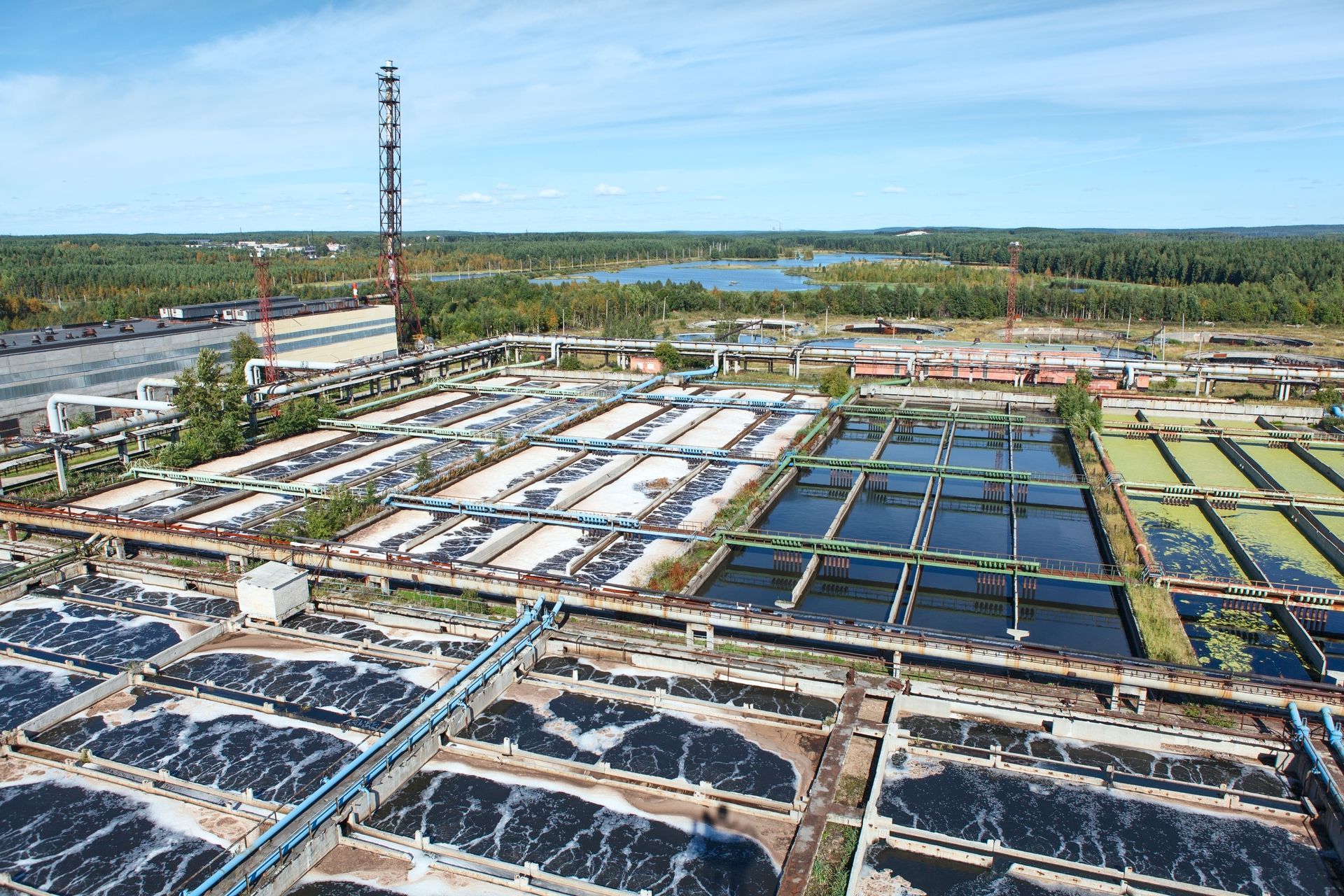 Aerial view of a wastewater treatment plant with rectangular basins and a tall smokestack, under a blue sky.