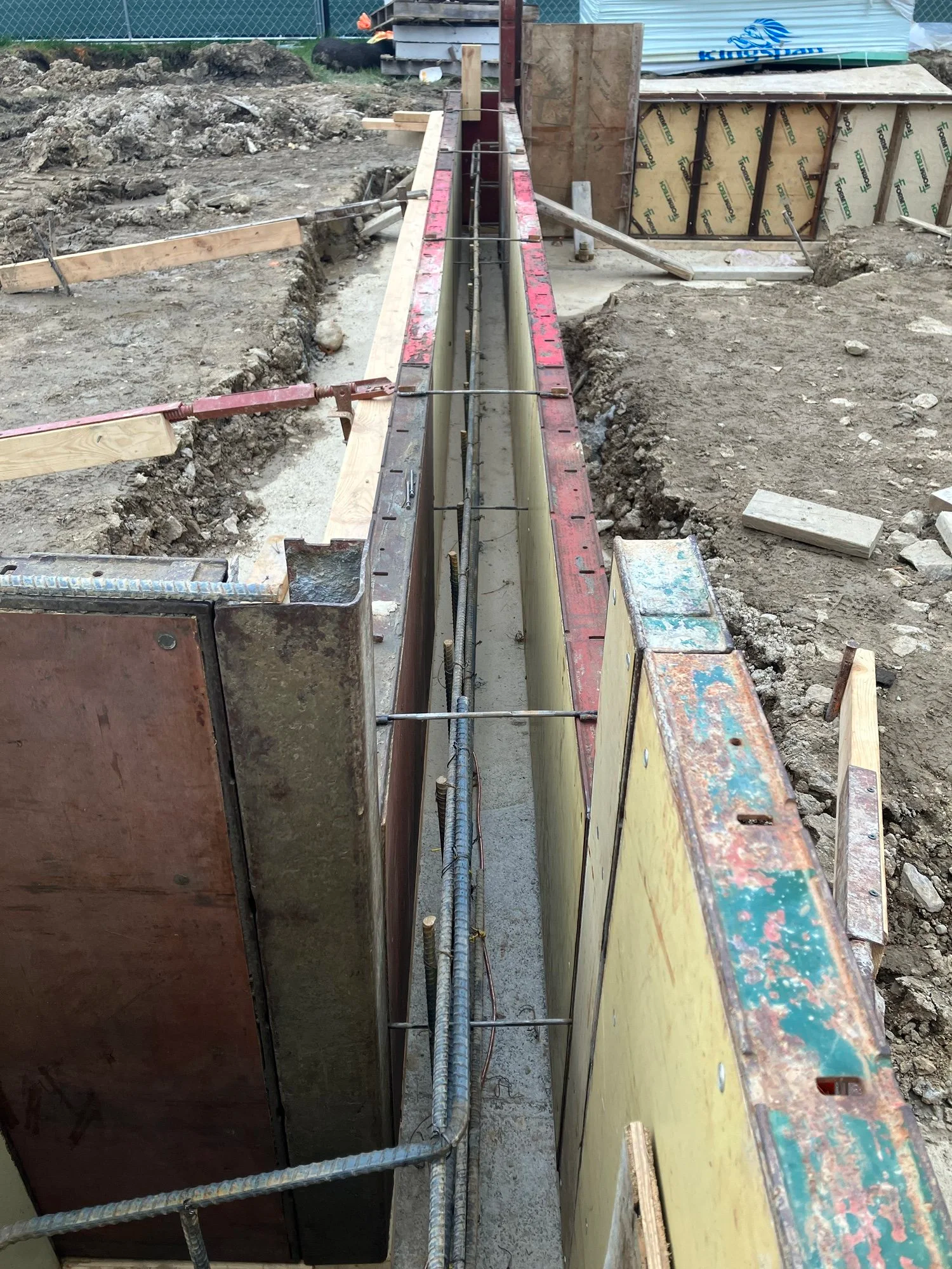 Construction site with a narrow concrete trench between formwork panels and metal rebar supports.