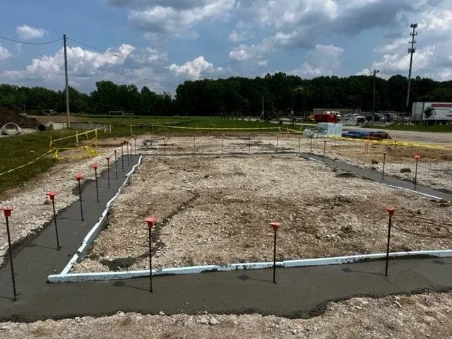 Construction site foundation with concrete footings and rebar stakes in a dirt lot under cloudy sky