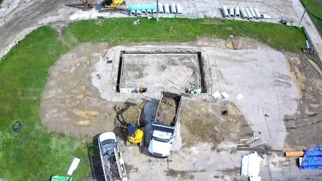 Aerial view of a construction site with an excavator and truck beside a large concrete foundation pit.