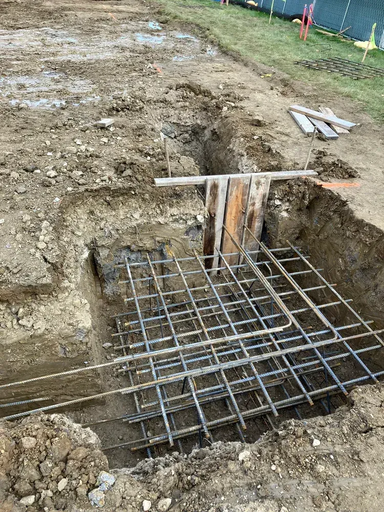 Construction trench with rebar grid and wooden formwork in muddy ground.