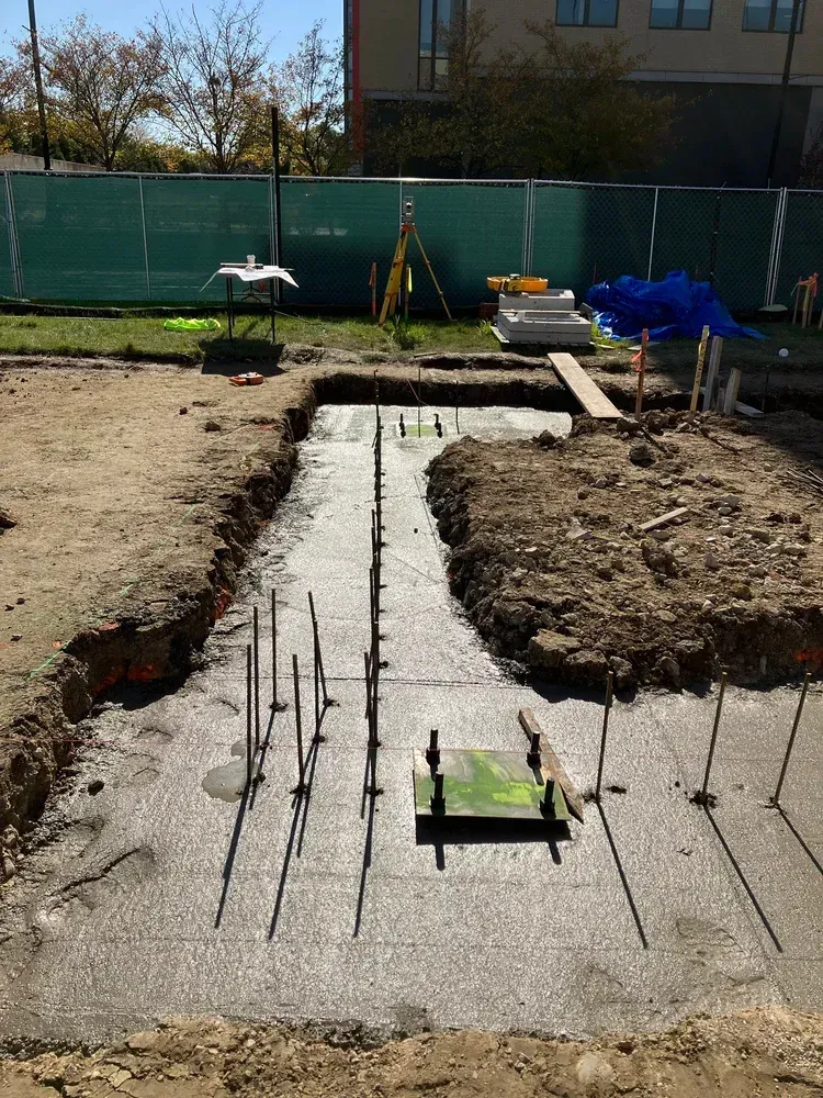 Construction site with concrete footing trench, exposed rebar, and dirt mounds beside a building.