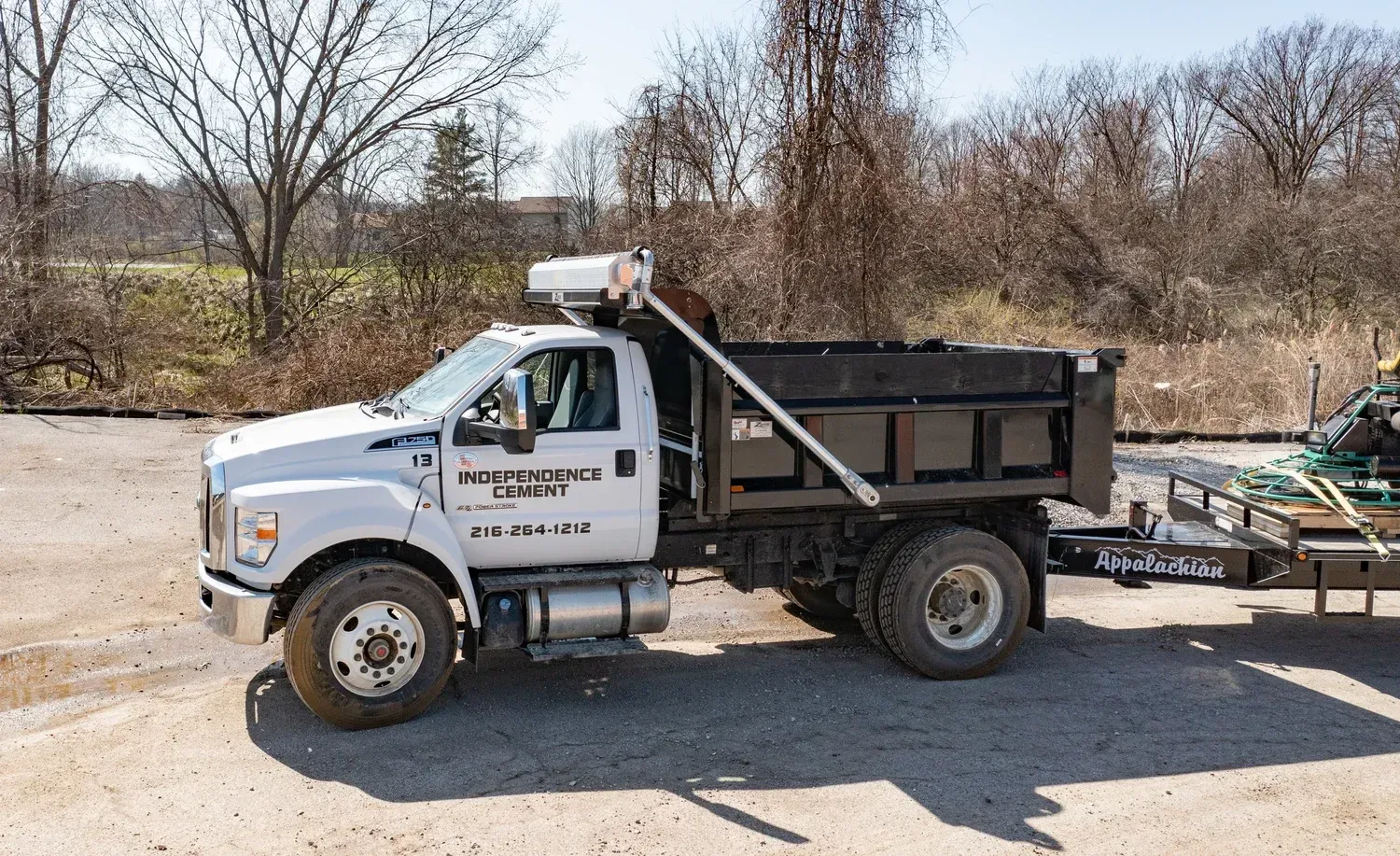 White utility truck with dump bed and trailer parked on a dirt road near trees and brush