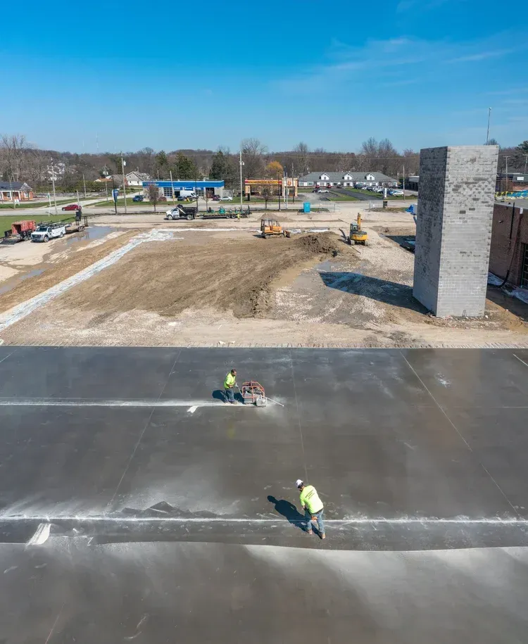 Road construction site with workers paving a large black roadway beside a dirt lot and unfinished building.