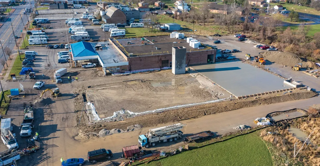 Aerial view of a construction site with a brick building, cleared lot, and parked vehicles on surrounding roads