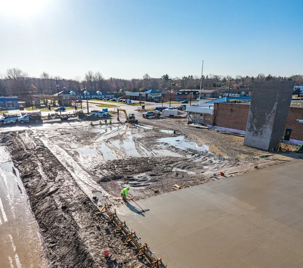 Snowy construction site with concrete foundation, muddy ground, and houses in the background