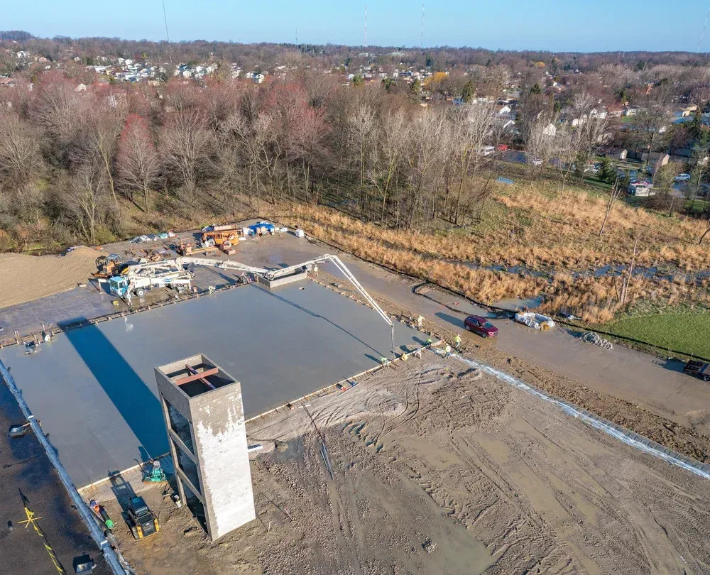 Aerial view of a construction site with a tall concrete shaft, paved lot, and trees in the background.