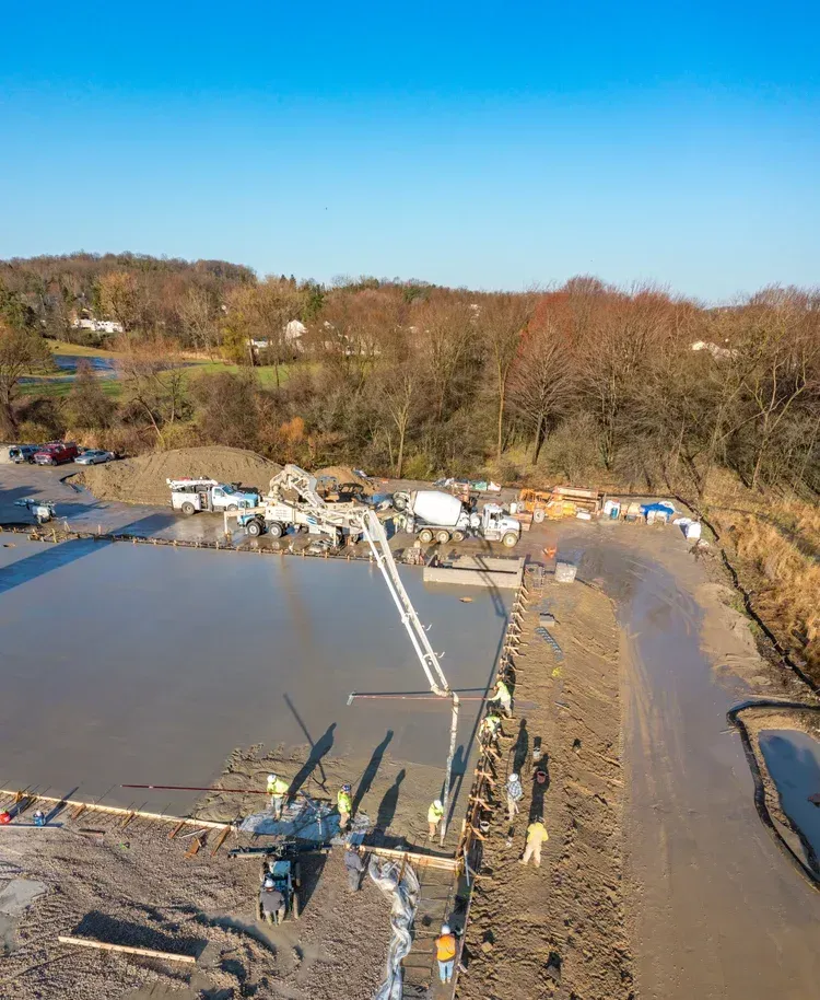Outdoor construction site with concrete foundation, trucks, and workers beside a muddy dirt road under a blue sky