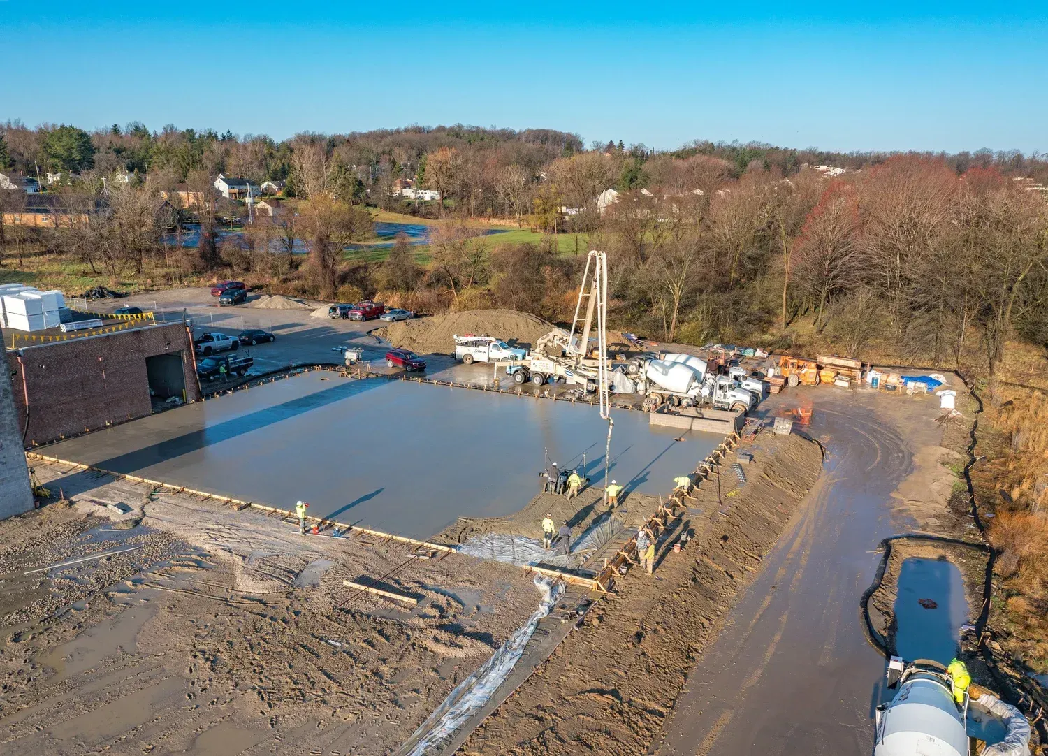Aerial view of a construction site with a concrete foundation, machinery, and surrounding wooded area under a clear sky