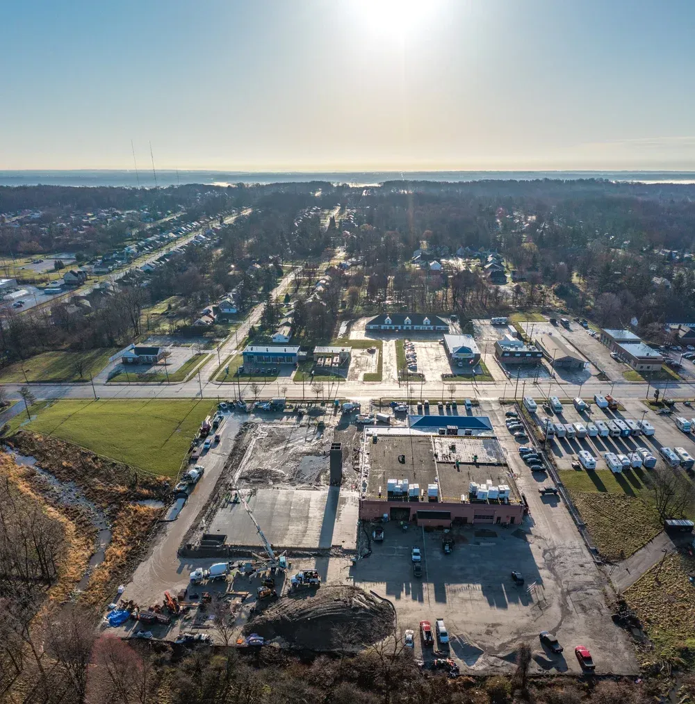 Aerial view of a commercial site with parking lots, roads, and wooded surroundings in bright sunlight