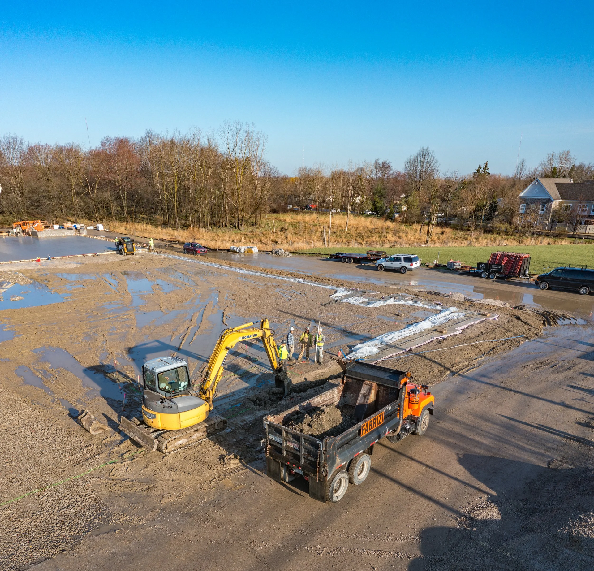 Construction site with excavator and dump truck on a muddy lot beside a road and trees