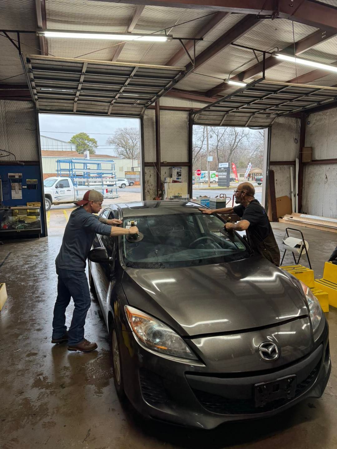 Two workers washing a gray sedan inside an auto shop bay