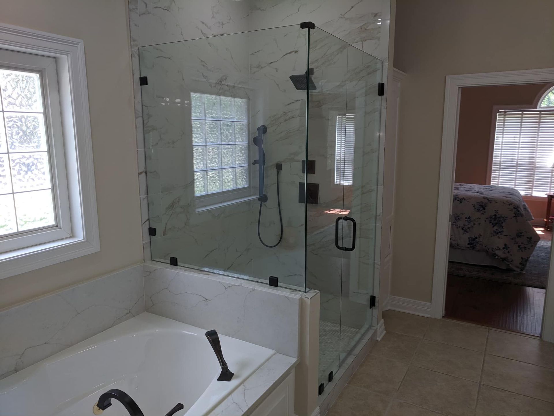 Modern bathroom with a glass shower, white tub, and window, leading into a bedroom.
