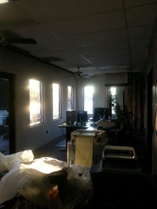 Dim office room with desks, sunlight through windows, and a potted plant near the back.