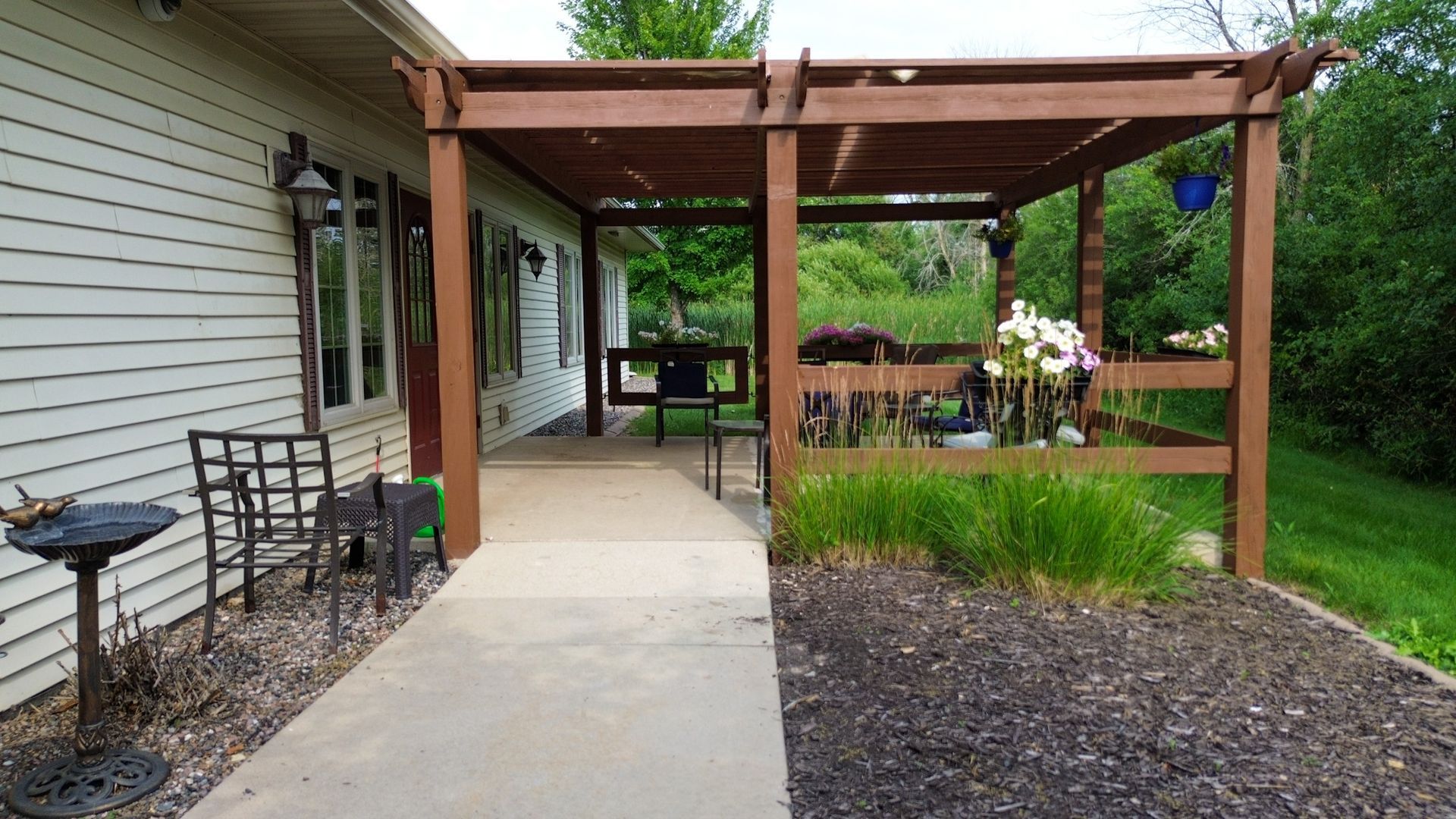 Brown pergola over a concrete walkway next to a house, with plants and furniture.