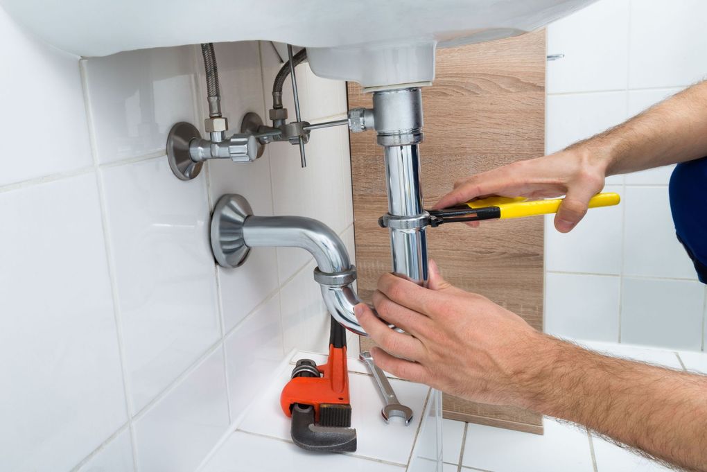 Plumber using a wrench to repair bathroom sink pipes; tools and tile visible.