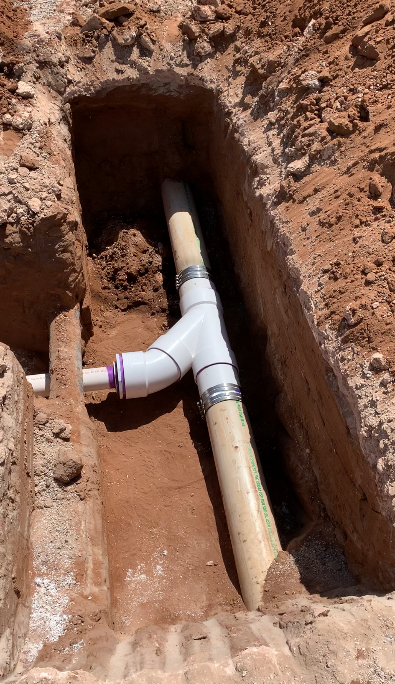 Trench with white PVC pipes, a tee fitting, and clamps in brown soil.