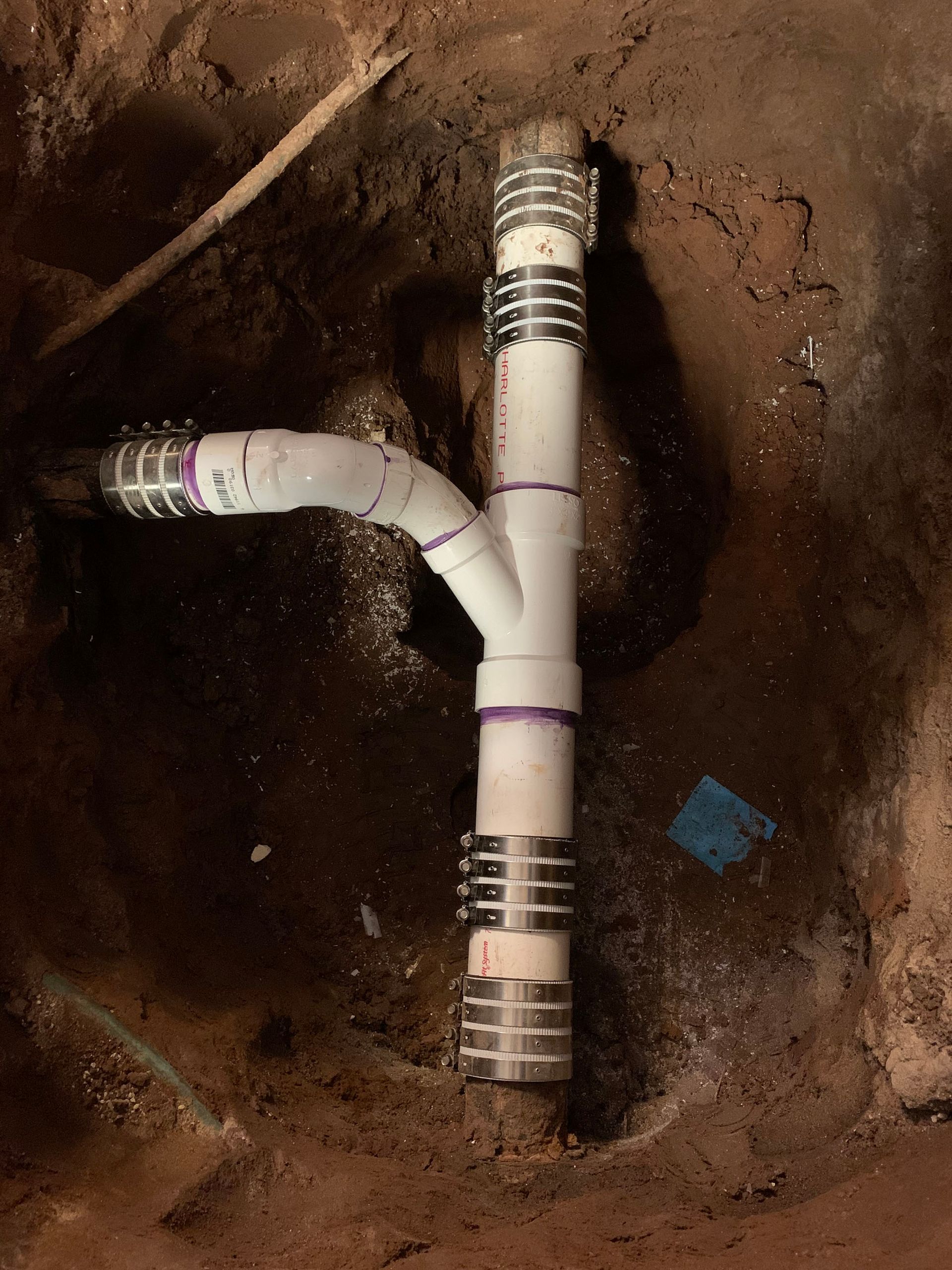 PVC plumbing pipes and fittings in a dirt pit. White pipes, metal clamps, purple fittings.