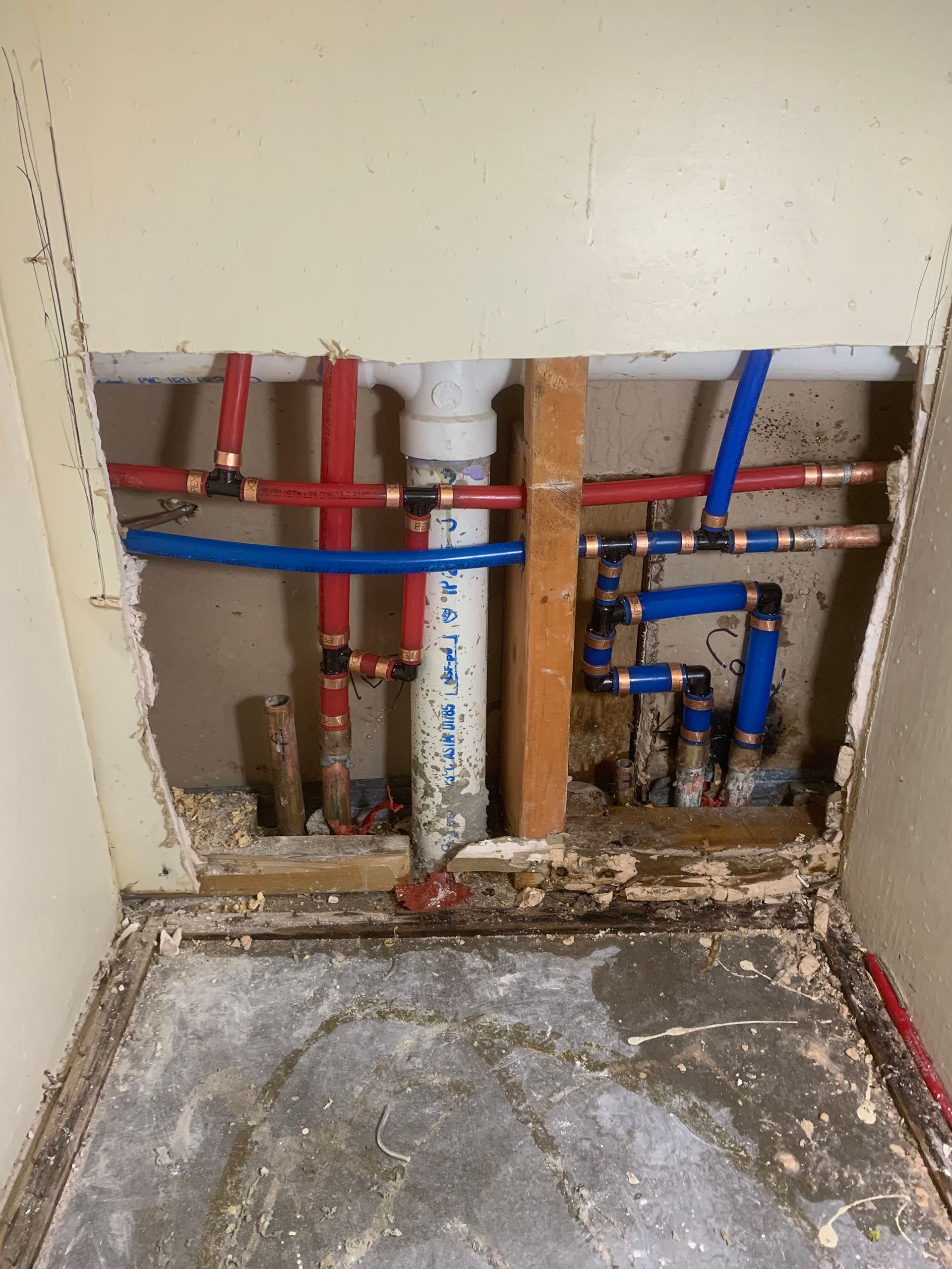 Pipes and plumbing exposed behind a damaged wall, red and blue water lines, white drain pipe.