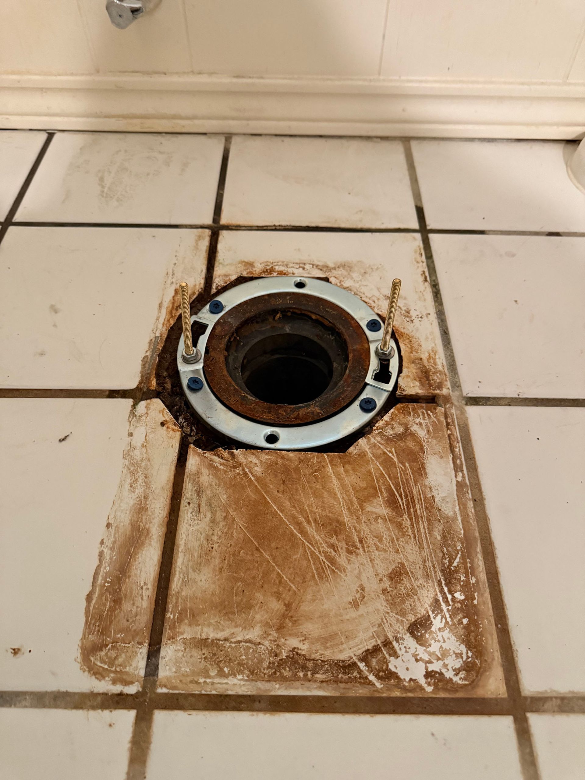 Toilet flange and bolts protruding from a hole in tiled floor, surrounded by old residue.