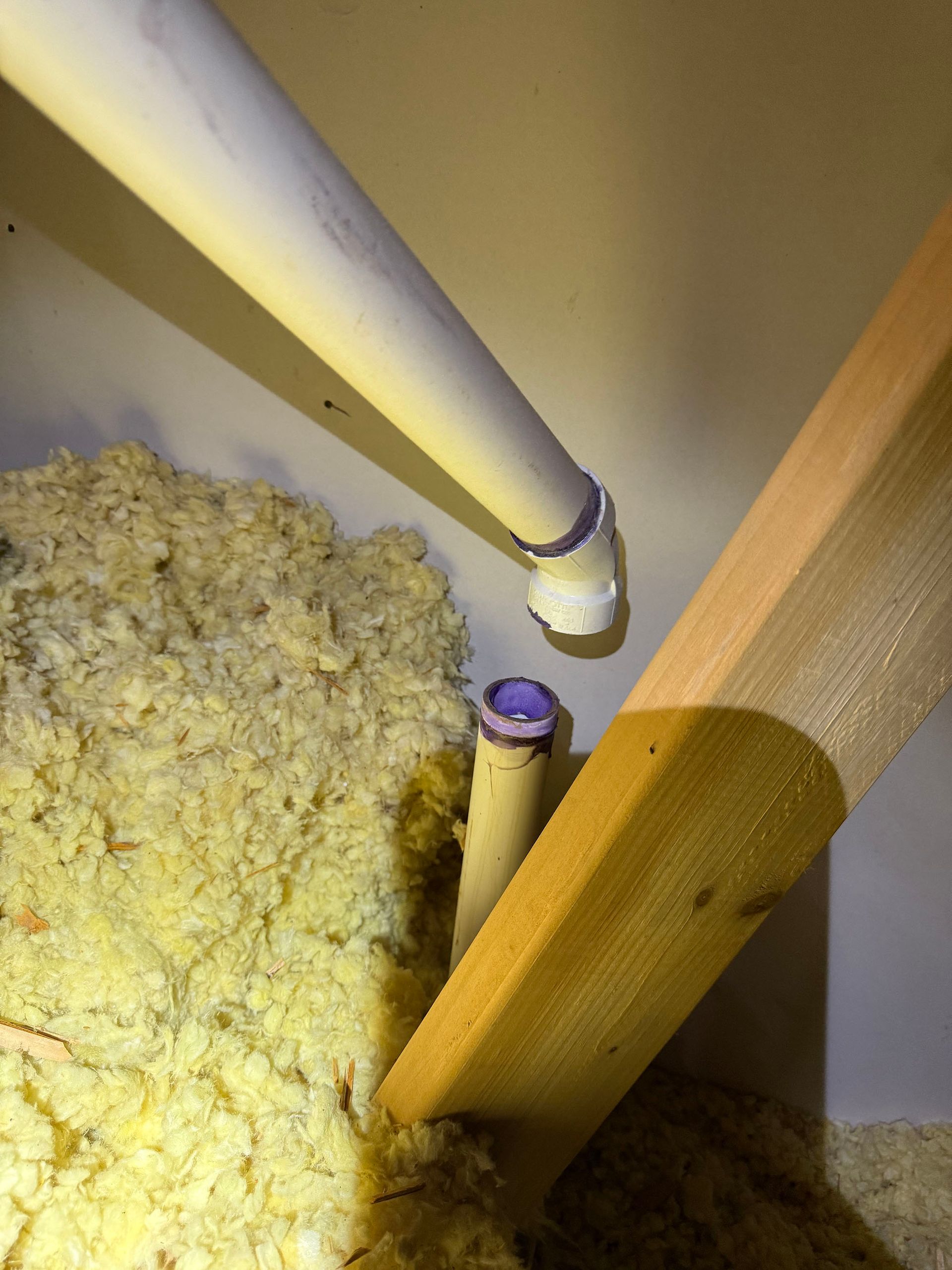 White PVC pipes in an attic with purple couplings and yellow insulation, angled near wood beams.