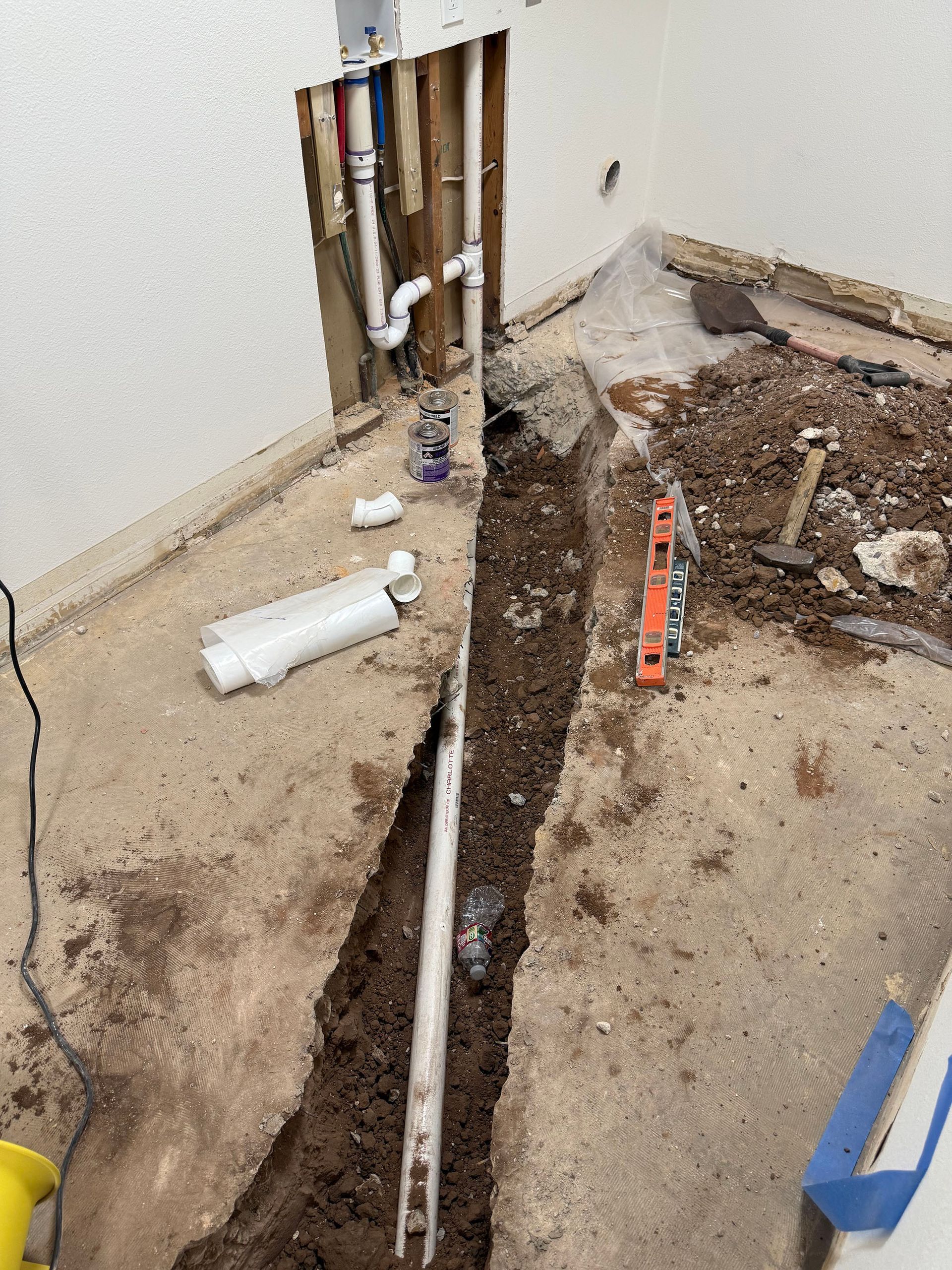 Trench dug in concrete floor, exposing plumbing pipes. White wall with exposed pipes, level and tools visible.