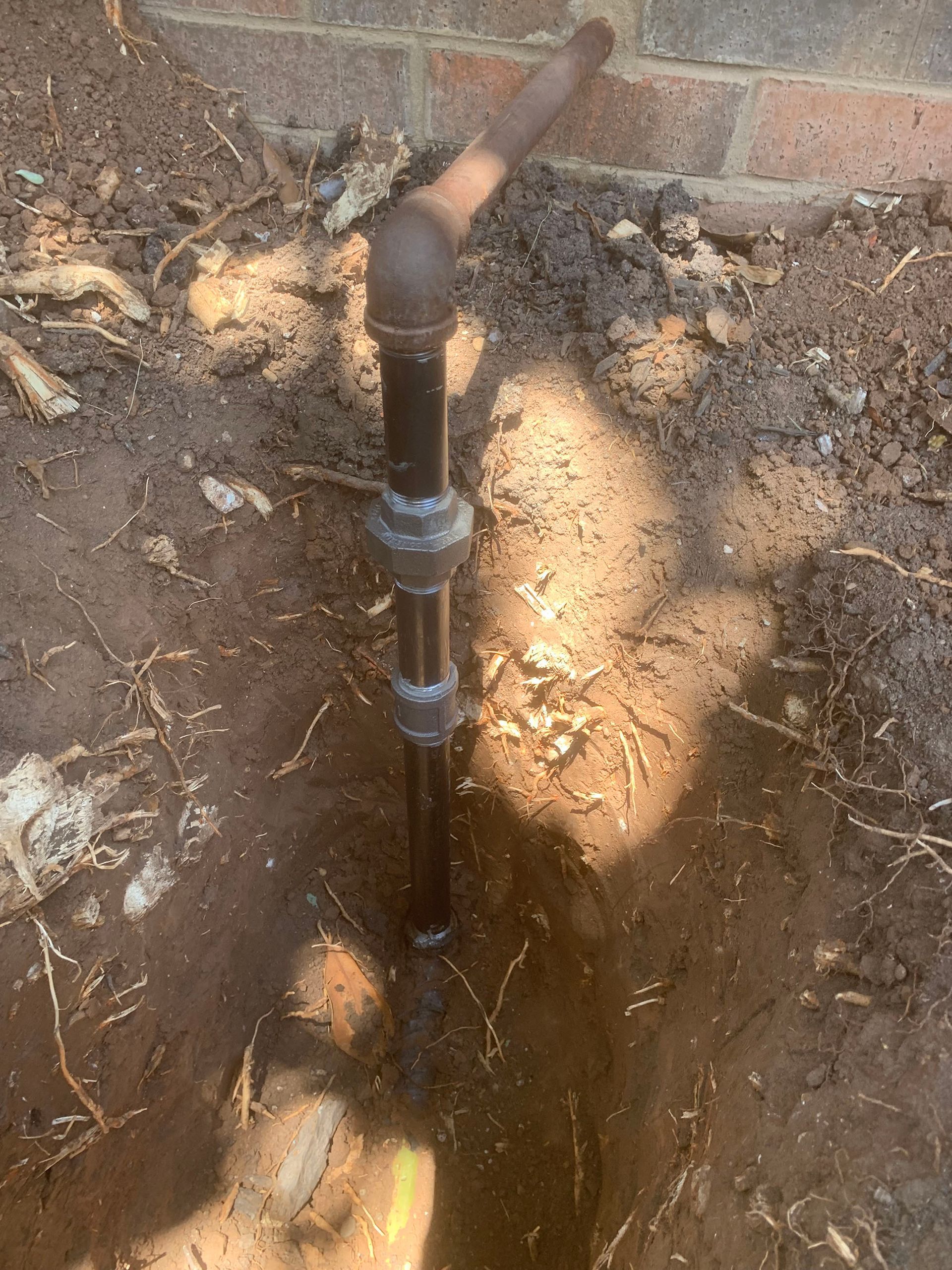 Black pipe in a trench near a brick wall, with exposed fittings.