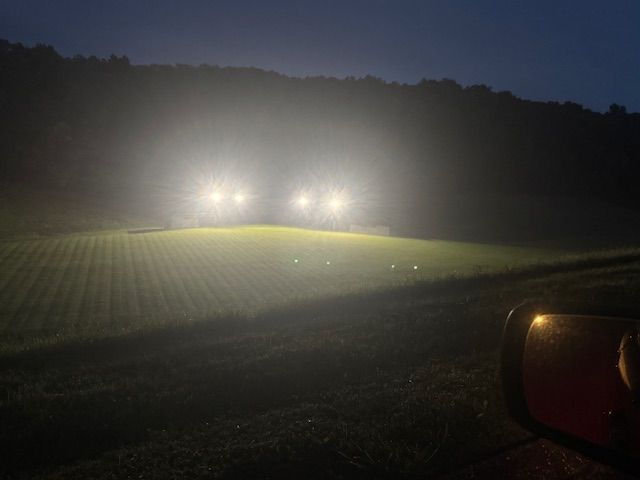 Field illuminated by bright lights at night, with trees in the background.  Viewed from a vehicle.