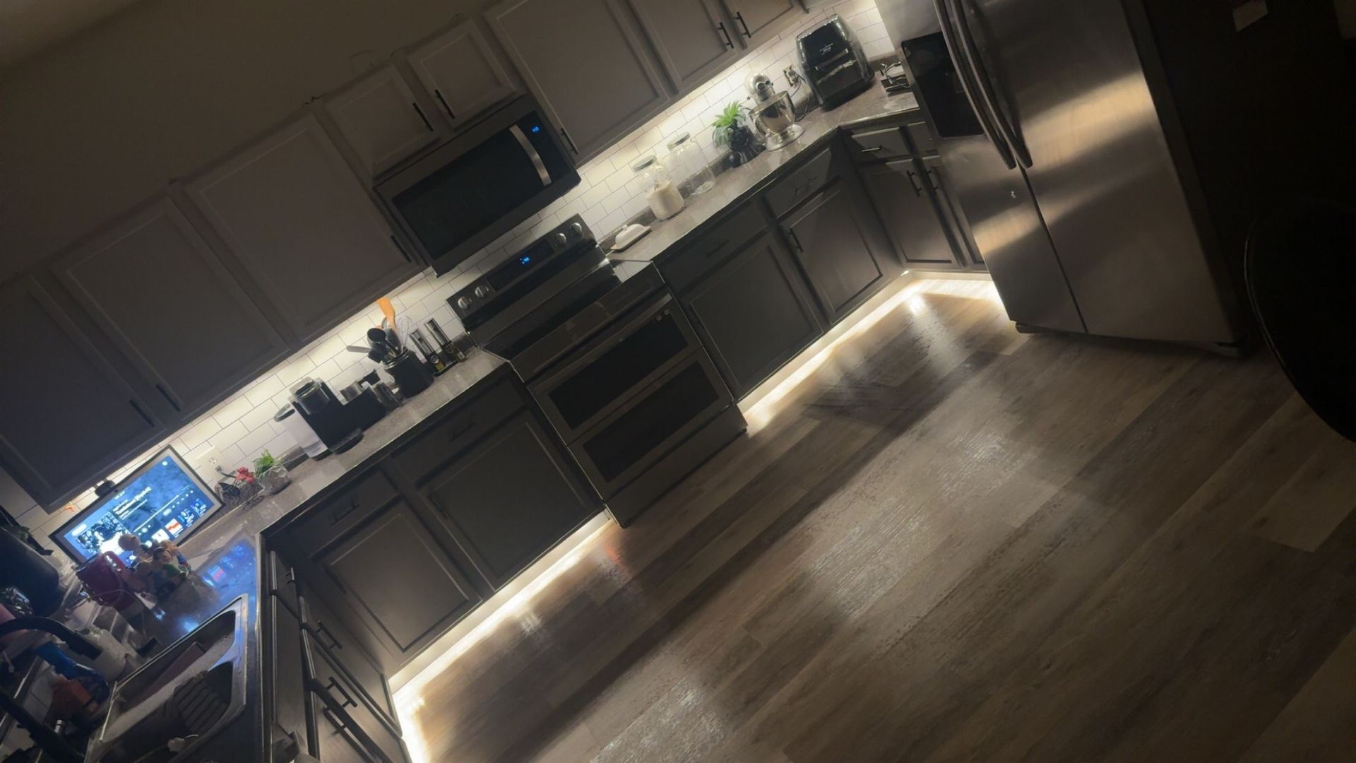 Kitchen with dark cabinets, white uppers, and under-cabinet lighting. Wooden floor reflects the light.