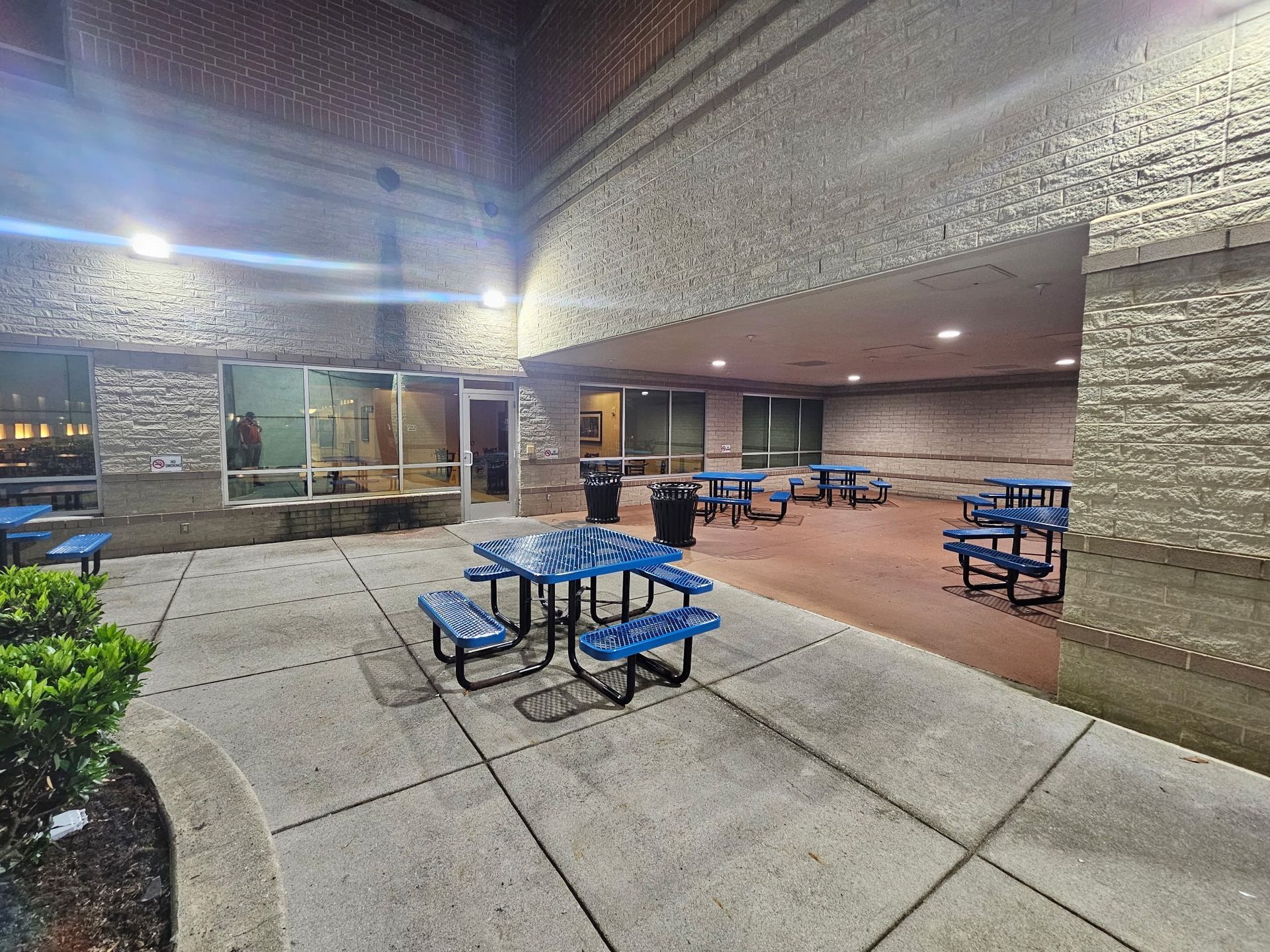 A covered outdoor patio with blue picnic tables, concrete flooring, and brick walls at night.
