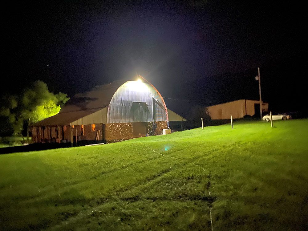 A brightly lit barn at night with a grassy hill in the foreground, and a white car in the distance