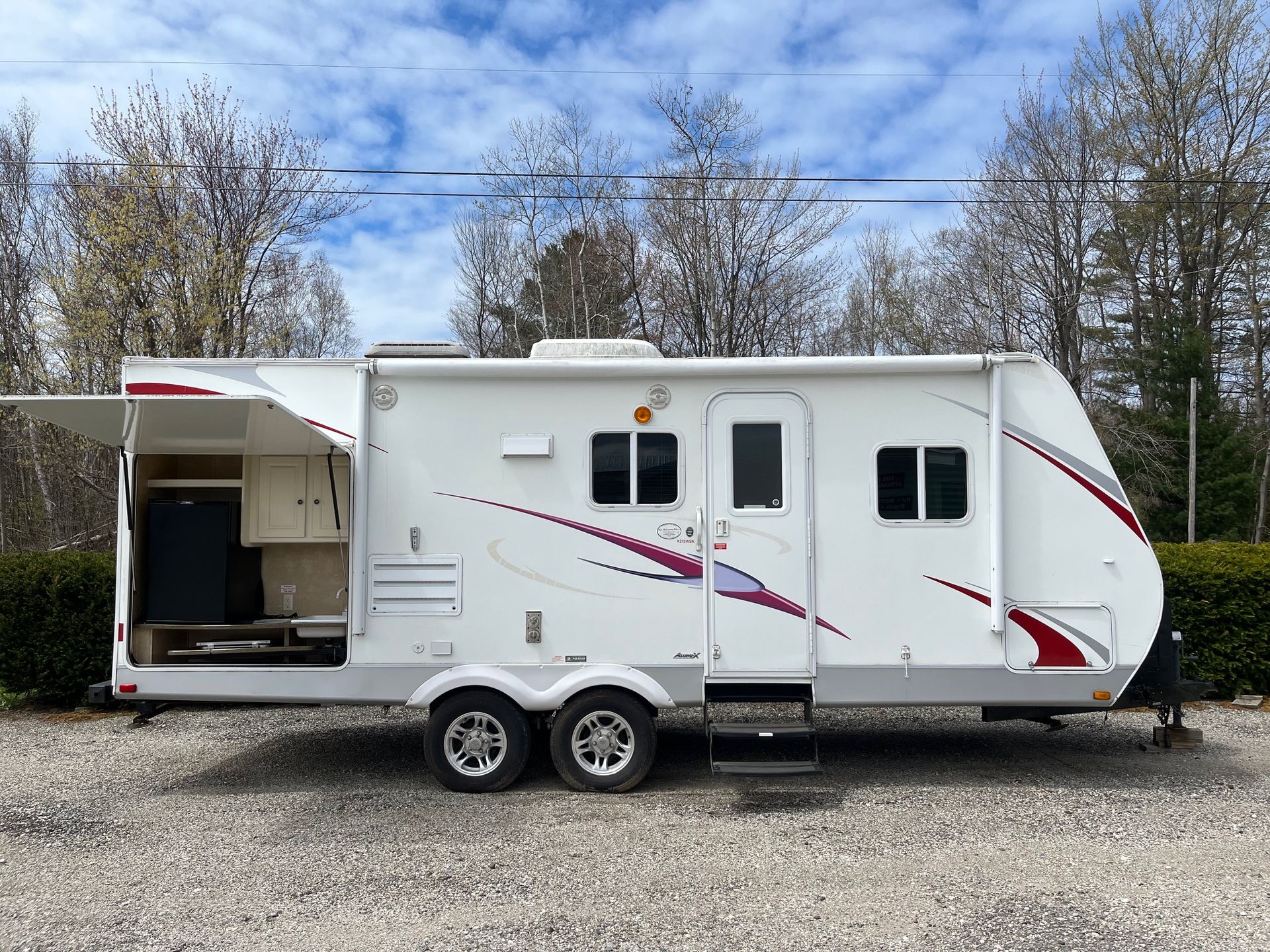 White dual-axle travel trailer with open rear cargo area parked on gravel under a cloudy sky
