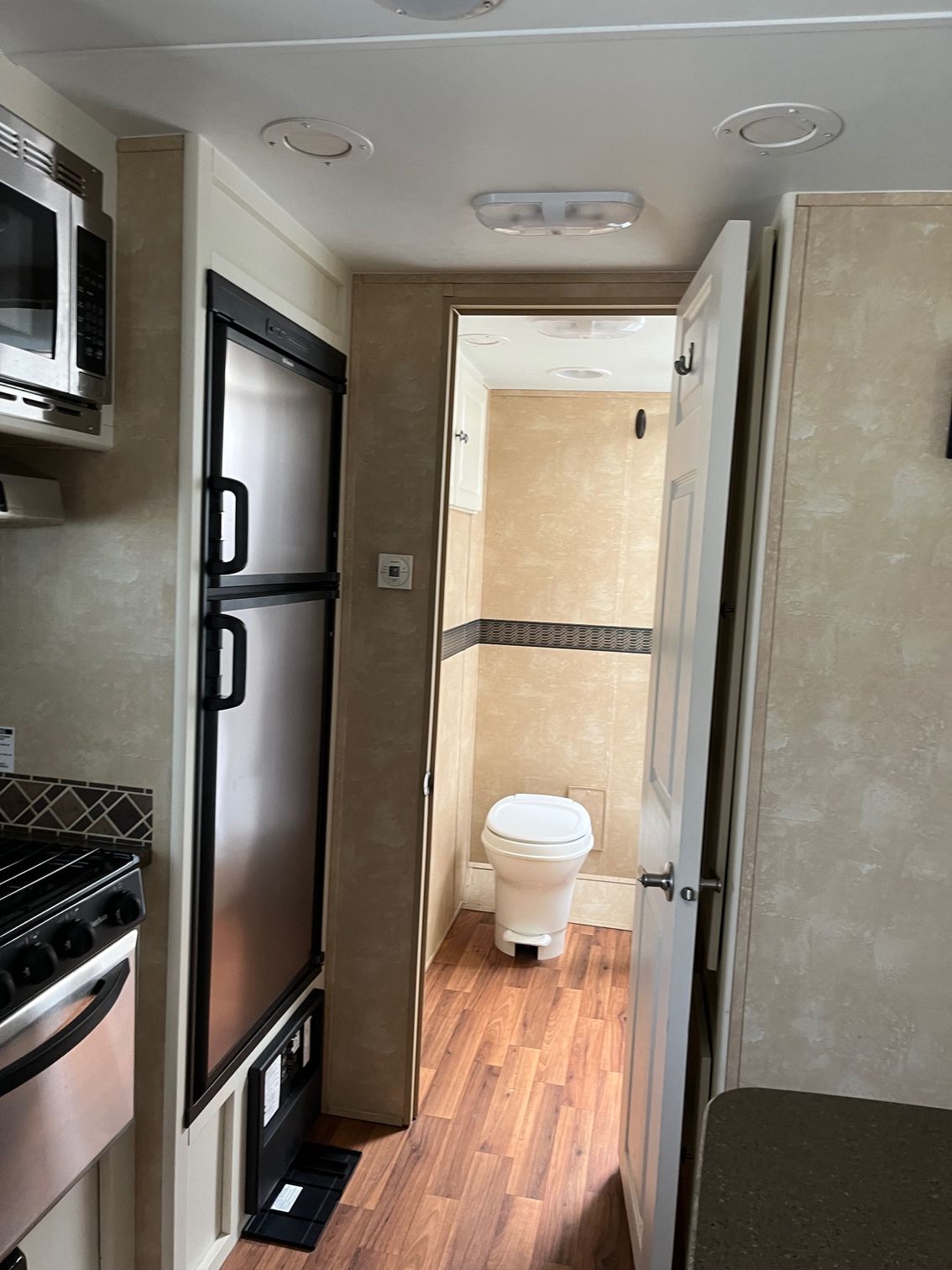 Narrow RV kitchen and hallway leading to a small bathroom with a toilet, wood flooring and beige walls