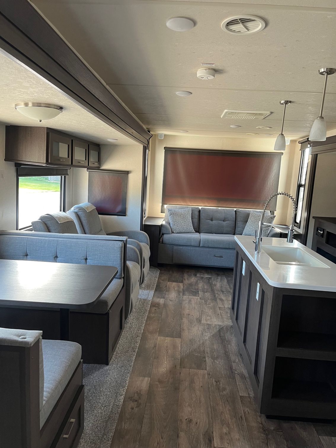 Modern RV interior with gray sofas, table, kitchen counter, and wood-look floor.