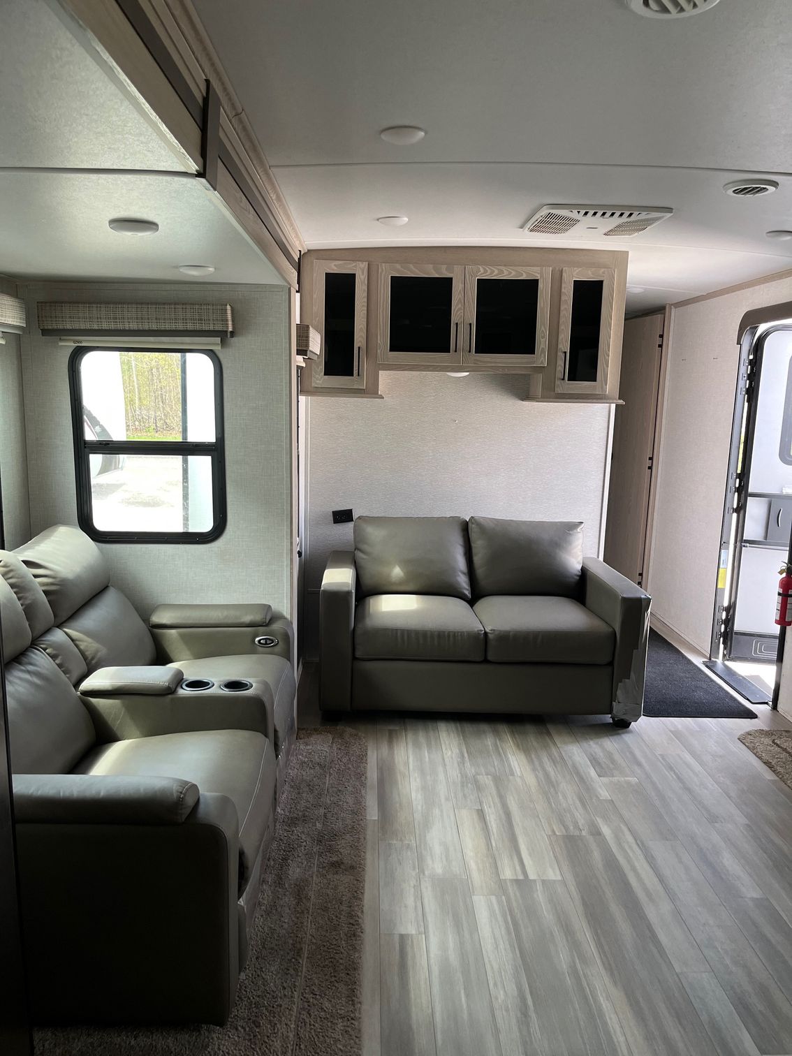 Small RV lounge with gray sofa, armchairs, wood-look floor, and windows along the walls.
