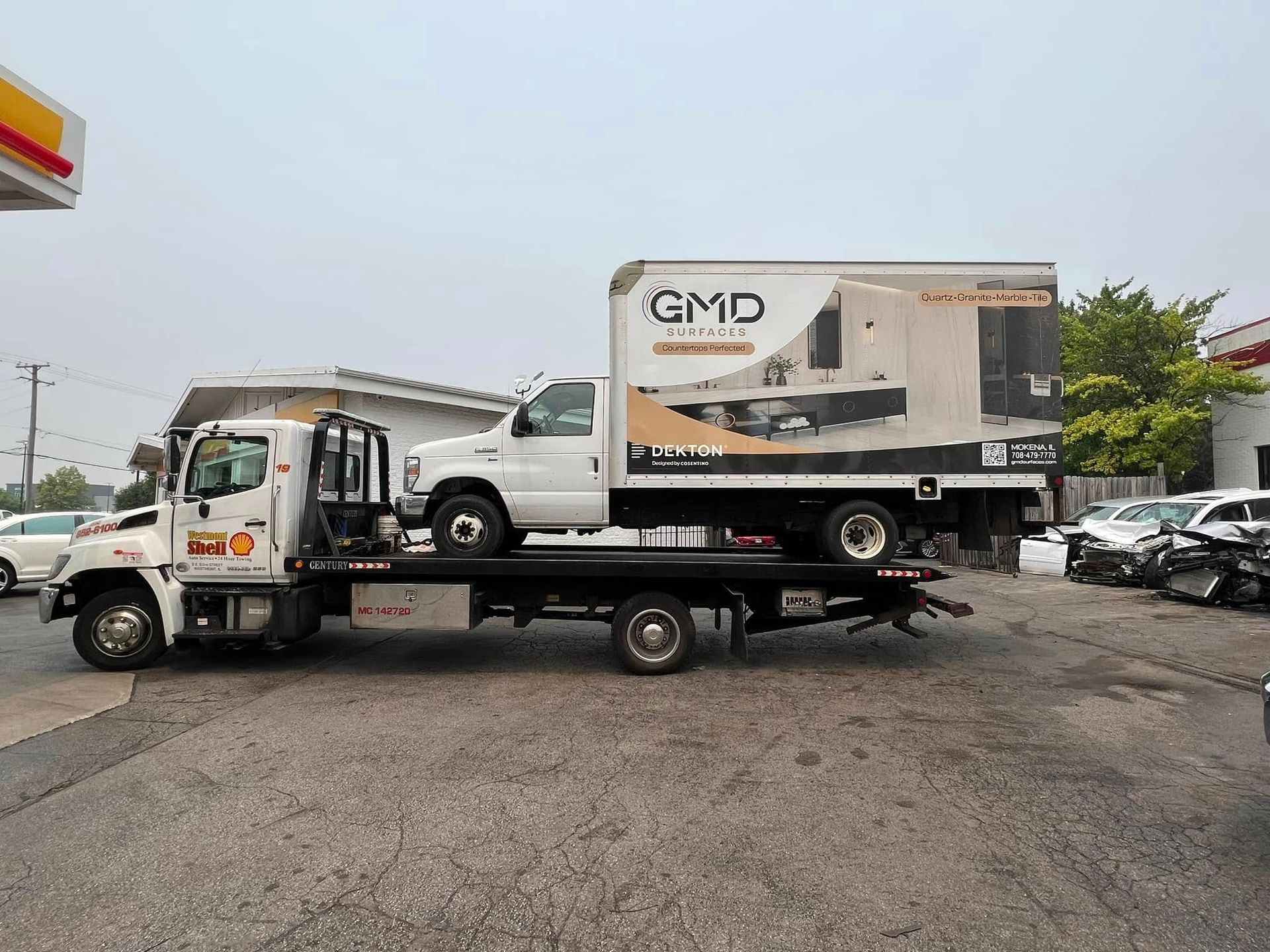 Tow truck carrying a white box truck in a gravel parking lot under cloudy sky