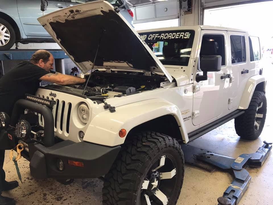 White Jeep Wrangler with hood open on a lift as a mechanic works in a garage