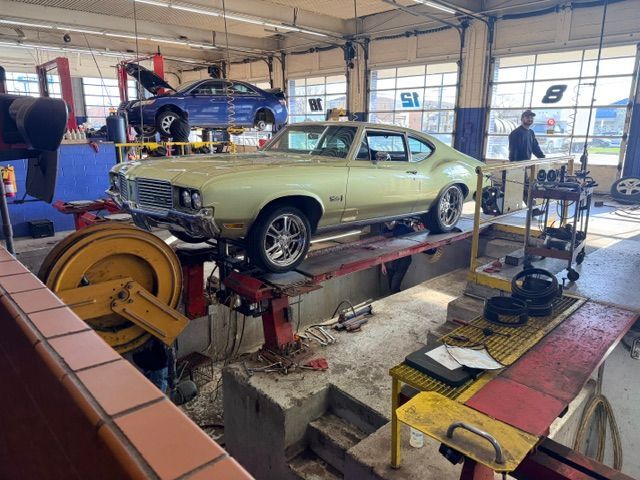 Classic green muscle car on a lift in an auto repair shop, with tools and another car in the background