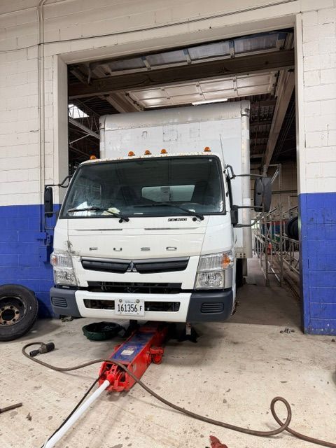 White truck in a garage bay, with a red floor jack under the front bumper and cables on the floor.